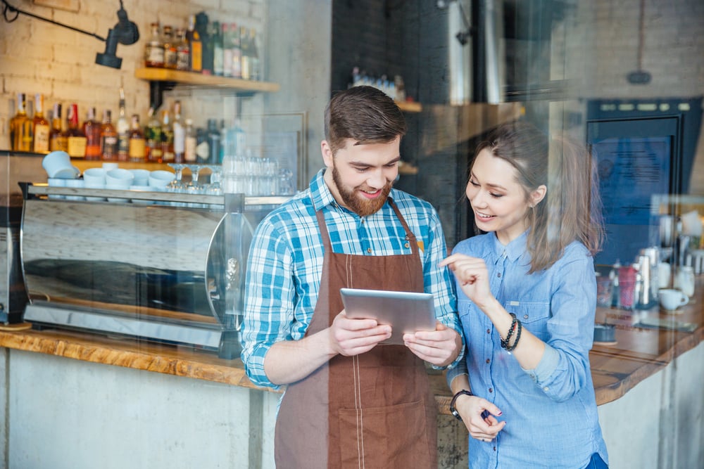 Sonriente camarero guapo sosteniendo la tableta y joven mujer bonita señalando en él en la cafetería